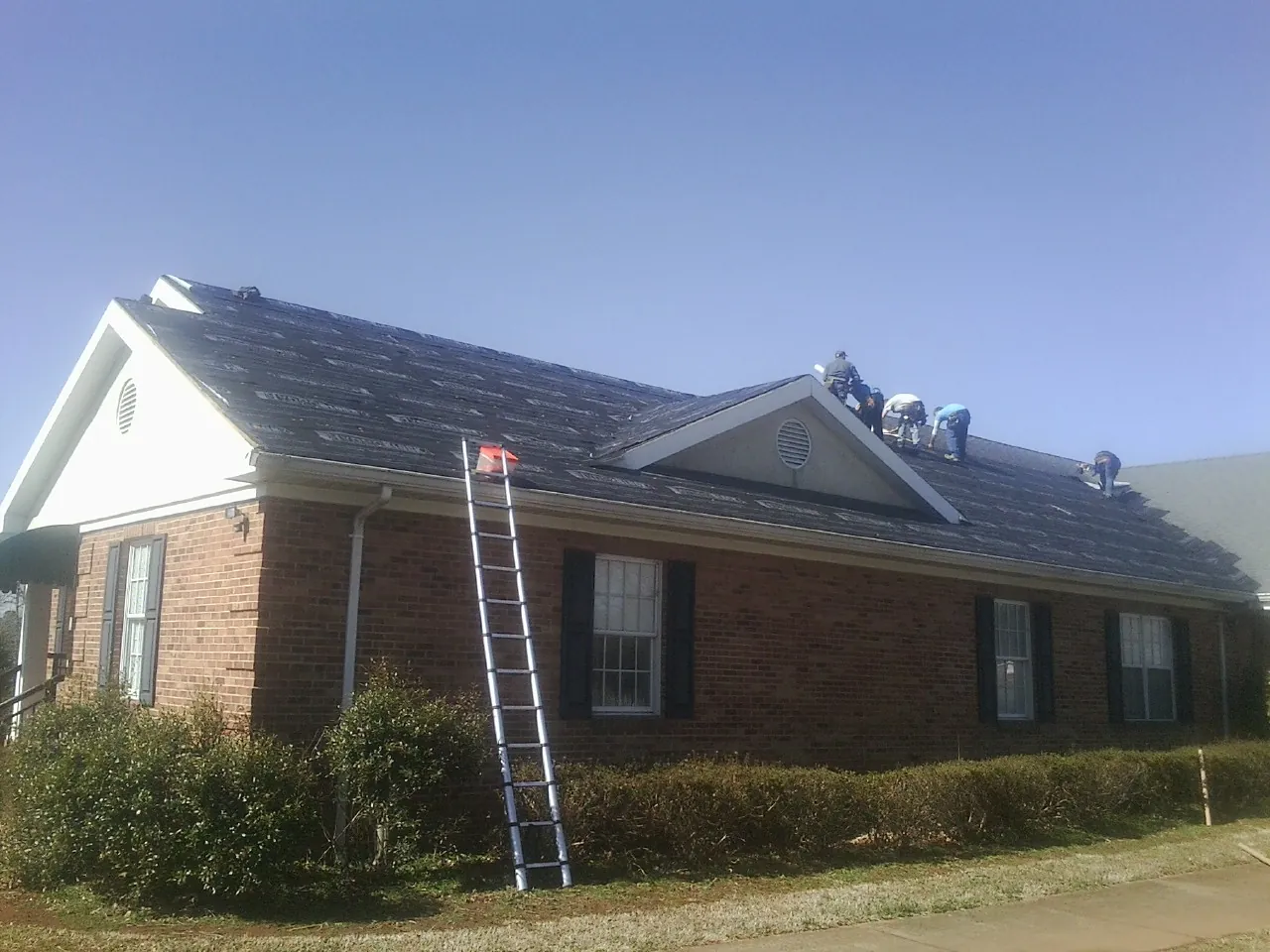 Crew tearing shingles off a brick ranch roof with a ladder set — Emergency Roof Repair work by our crew in Mamou