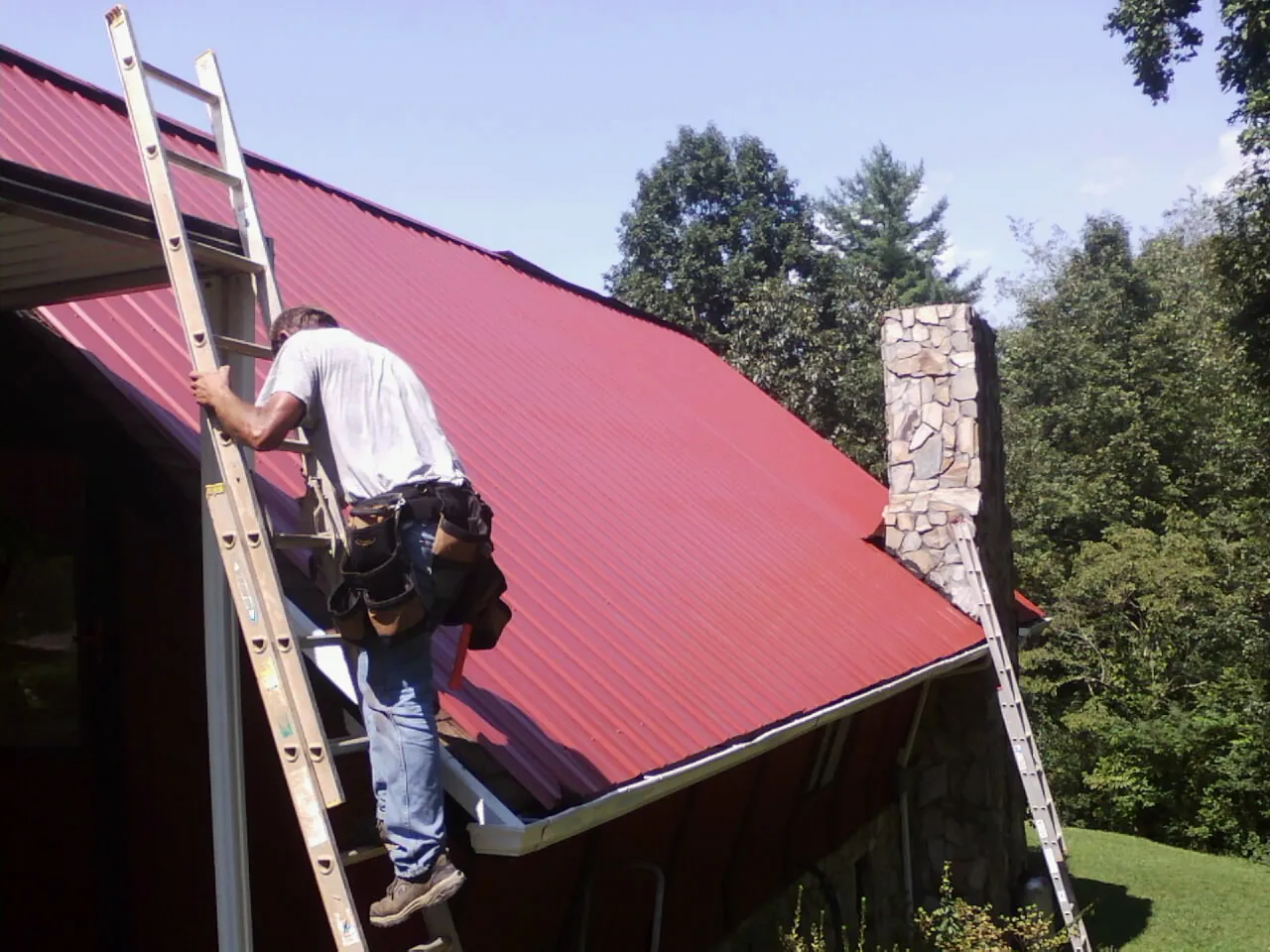 Roofer on a ladder beside a red metal roof and stone chimney — Soffit & Fascia Repair by Roofing Brothers in Mamou